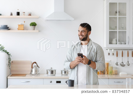 Happy adult caucasian guy with smartphone and cup of coffee look at empty space at kitchen interior Happy adult caucasian guy with smartphone and cup of coffee look at empty space at kitchen interior 83734857