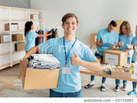 Portrait of young male volunteer holding cardboard box for donation, smiling at camera and showing thumbs up 83734861