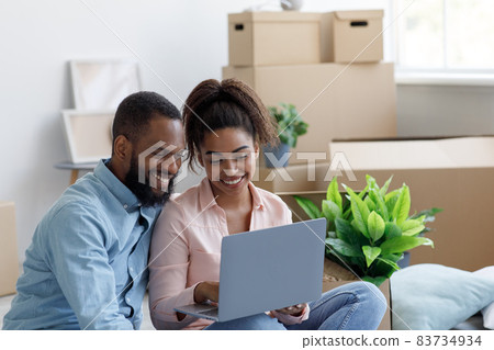 Smiling young black man and lady in new apartment among boxes, planning interior, couple using laptop Smiling young black man and lady in new apartment among boxes, planning interior, couple using laptop 83734934