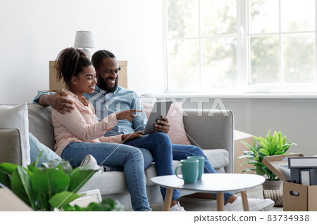 Satisfied young african american man and woman sit on sofa, rest among boxes and plants after moving Satisfied young african american man and woman sit on sofa, rest among boxes and plants after moving 83734938