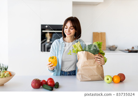 Young lady unpacking grocery bag after shopping, putting vegetables on table in kitchen interior at home, copy spsce Young lady unpacking grocery bag after shopping, putting vegetables on table in kitchen interior at home, copy spsce 83735024