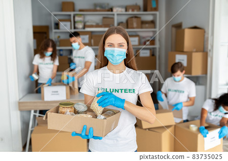 Young female volunteer wearing volunteer uniform and medical mask, holding box with canned food and grains Young female volunteer wearing volunteer uniform and medical mask, holding box with canned food and grains 83735025