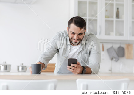 Cheerful mature european man with cup of coffee reads news and checks social networks at kitchen Cheerful mature european man with cup of coffee reads news and checks social networks at kitchen 83735026