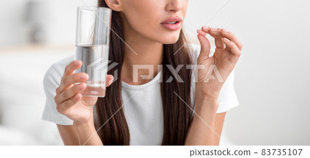 Caucasian young woman holding pill and glass of water in bedroom interior, panorama, cropped 83735107