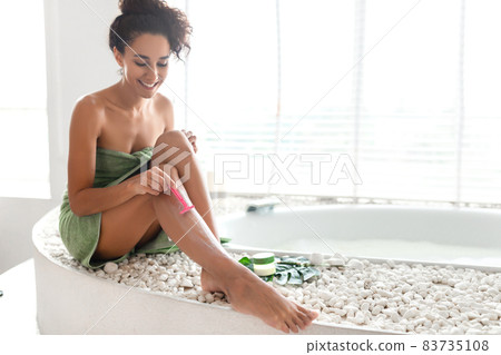 Lovely young woman shaving legs with razor, sitting in towel on bathtub, caring for body in modern bathroom, copy space Lovely young woman shaving legs with razor, sitting in towel on bathtub, caring for body in modern bathroom, copy space 83735108