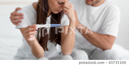 Cropped disappointed sad young caucasian man support his wife, hold pregnancy test, sit on bed in bedroom 83735109