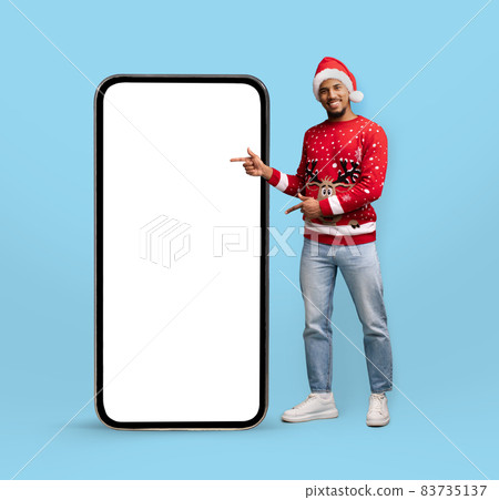 Cheerful black guy wearing Santa hat, pointing at cellphone, mockup 83735137