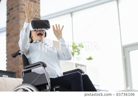 Woman sitting in wheelchair wearing virtual reality glasses and waving her hands Woman sitting in wheelchair wearing virtual reality glasses and waving her hands 83736106
