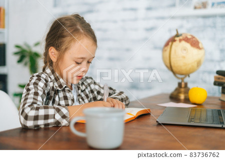 Side view of focused primary child school girl doing writing homework sitting at table with laptop and paper notebook at home. Side view of focused primary child school girl doing writing homework sitting at table with laptop and paper notebook at home. 83736762