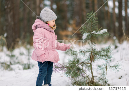 Happy winter concept. A little girl in a pink jacket and gray cap is playing with pine tree branch and smiling. Happy girl in winter snowy forest. Copy space. 83737615