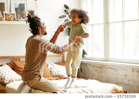 Happy african american father and son jumping on bed and laughing while playing together at home Happy african american father and son jumping on bed and laughing while playing together at home 83738509
