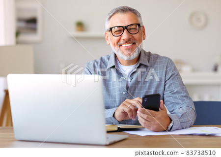 Smiling senior man using mobile phone while sitting at his cozy workplace with laptop at home Smiling senior man using mobile phone while sitting at his cozy workplace with laptop at home 83738510