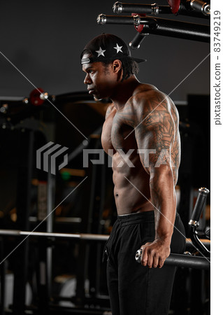 Strong and muscular dark skin man trains on modern equipment in gym. Portrait of muscular pumped up fitness trainer Strong and muscular dark skin man trains on modern equipment in gym. Portrait of muscular pumped up fitness trainer 83749219
