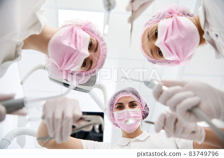Bottom view of female dentists and dental asistant in pink protective mask holding sterile tools for a dental examination and leaning towards a patient . Focus on dental instruments Bottom view of female dentists and dental asistant in pink protective mask holding sterile tools for a dental examination and leaning towards a patient . Focus on dental instruments 83749796