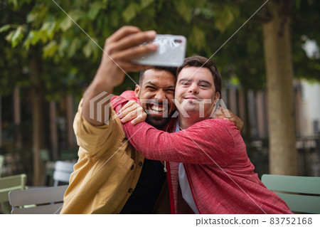 Young man with Down syndrome and his mentoring friend sitting and taking selfie outdoors in cafe 83752168