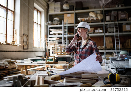 Female engineer holding blueprints and making phone call indoors in carpentry workshop. 83752213