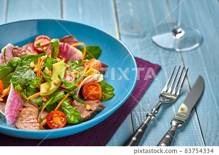 Spicy beef slices meat salad with tomatoes, parsley, radish and salad leaves spinach in a blue plate on a wooden table. 83754334