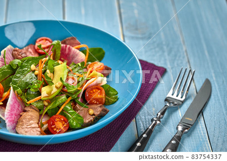 Spicy beef slices meat salad with tomatoes, parsley, radish and salad leaves spinach in a blue plate on a wooden table. 83754337