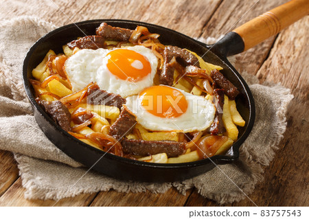 Chorrillana, a Chilean popular dish made with fries, and topped with beef onions and eggs close up in the pan. Horizontal 83757543