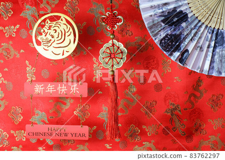 Golden chinese tiger symbol, chinese bamboo paper fan and fortune hunger from lucky coins on red fabric with birds and dragons. Chinese New Year of the Tiger 2022. Translation: happy new year Golden chinese tiger symbol, chinese bamboo paper fan and fortune hunger from lucky coins on red fabric with birds and dragons. Chinese New Year of the Tiger 2022. Translation: happy new year 83762297