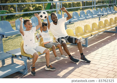 Black father and two his multiracional sons watching football game on stadium 83762325