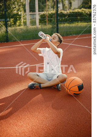 Multiracial boy sitting on a basketball court with a bottle of water Multiracial boy sitting on a basketball court with a bottle of water 83762376