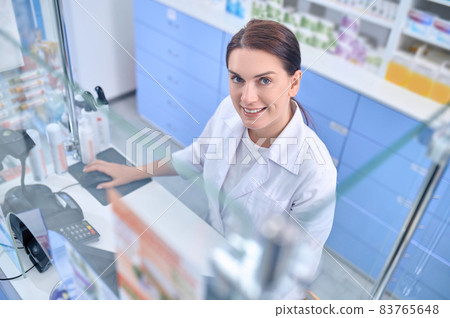 Joyful woman pharmacist behind cash register Joyful woman pharmacist behind cash register 83765648