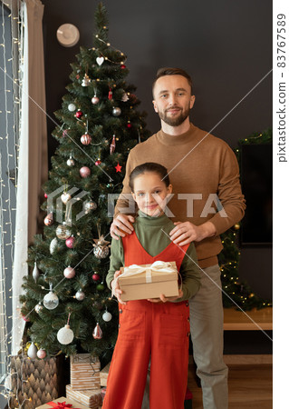 Portrait of handsome middle-aged father in sweater embracing cute daughter with gift box against Christmas tree Portrait of handsome middle-aged father in sweater embracing cute daughter with gift box against Christmas tree 83767589