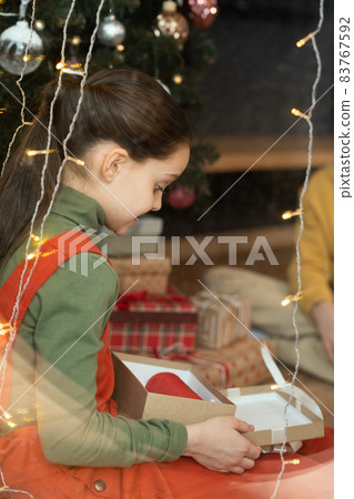 Brunette girl in turtleneck sitting at Christmas tree and looking at red portable speaker in box Brunette girl in turtleneck sitting at Christmas tree and looking at red portable speaker in box 83767592