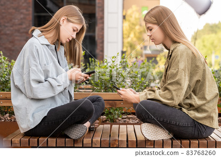 Twin girls sitting on bench in front of one another 83768260