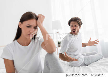 Aggressive evil millennial caucasian husband with stubble shouts and swears at offended sad depressed wife in bedroom Aggressive evil millennial caucasian husband with stubble shouts and swears at offended sad depressed wife in bedroom 83768446