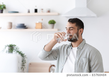 Middle aged european attractive guy with beard drinks water from glass on minimalist kitchen interior 83768484