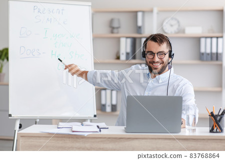 Smiling middle aged caucasian guy teacher in glasses, headphones shows at blackboard with english rules 83768864