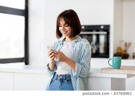Online ordering food. Excited young lady typing on smartphone in modern kitchen interior, free space Online ordering food. Excited young lady typing on smartphone in modern kitchen interior, free space 83768962