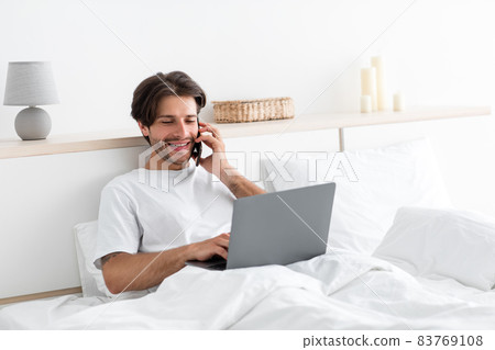 Cheerful attractive millennial european man works on laptop, speaks on phone with client on bed 83769108