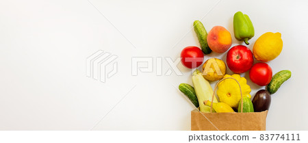 A paper shopping bag with vegetables and fruits, tomato, cucumber, squash, pepper, lemon, eggplant, zucchini, banana, apple, peach on white background A paper shopping bag with vegetables and fruits, tomato, cucumber, squash, pepper, lemon, eggplant, zucchini, banana, apple, peach on white background 83774111