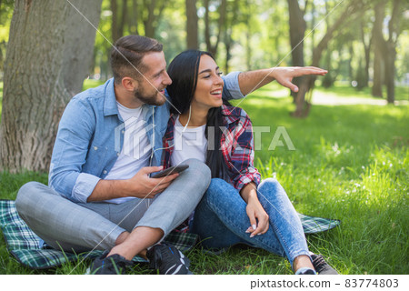 Couple in love are resting in the park and hugging. Man is showing something to girl. Love couple sitting on a plaid blanket in the park and use the phone, handsome guy and girl 83774803