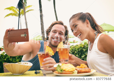 Selfie photo couple taking picture with phone at Hawaii beach bar on summer vacation. Happy Asian woman and man toasting mai tai drinks at bar having fun. Friends on travel holidays. 83777688