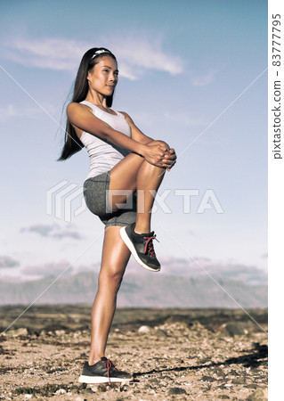 Leg stretching exercise fitness runner woman doing running warm-up, hamstring muscles stretch standing wtih single knee to chest stretch. Female athlete preparing legs for cardio workout. 83777795