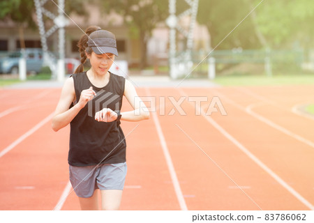 Portrait of young athlete runner woman running in the running track and she looking to her smart watch. Running track is a rubberized artificial running surface for track and field athletics. Portrait of young athlete runner woman running in the running track and she looking to her smart watch. Running track is a rubberized artificial running surface for track and field athletics. 83786062