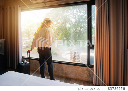 Portrait of tourist woman standing nearly window with her luggage in hotel bedroom after check-in. Conceptual of travel and vacation. 83786256