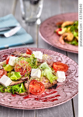 Healthy mixed Greek salad served on a pink plate with silver fork containing crisp leafy greens, microgreen, feta, onion, tomato and sliced beef 83786840