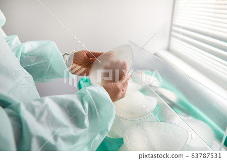 Breast prothesis in surgeon hands on background of medical samples of breast silicone implants of various sizes for breast augmentation aesthetic plastic surgery after resection due to cancer disease 83787531