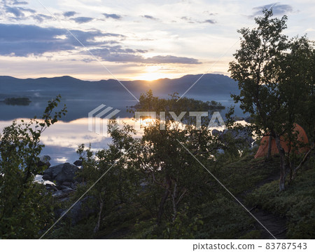Beautiful pink golden light sunrise at lake dam on river Lulealven in Saltoluokta in Sweden Lapland. Blue mountains, birch trees and clouds, sun reflecting on water surface. Summer northern landscape 83787543