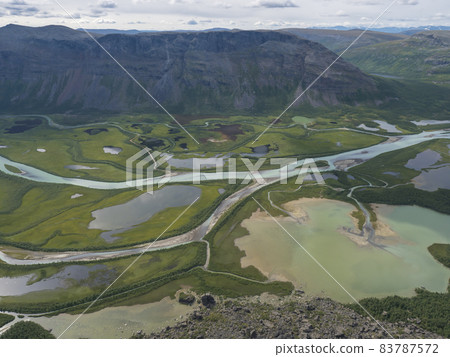 Aerial scenic view from Skierffe rock summit on glacial Rapadalen river delta valley at Sarek national park with meanders, lakes, mountains and birch trees. Summer day landscape Sweden Lapland Aerial scenic view from Skierffe rock summit on glacial Rapadalen river delta valley at Sarek national park with meanders, lakes, mountains and birch trees. Summer day landscape Sweden Lapland 83787572
