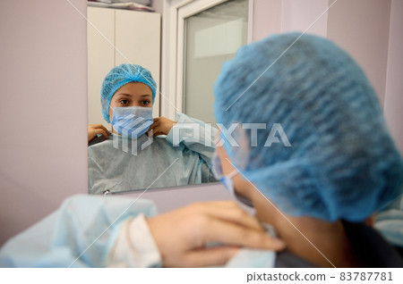 View from the back of a medical professional, doctor, nurse, surgeon in protective workwear and medical mask looking at his mirror reflection in dressing room of medical clinic View from the back of a medical professional, doctor, nurse, surgeon in protective workwear and medical mask looking at his mirror reflection in dressing room of medical clinic 83787781