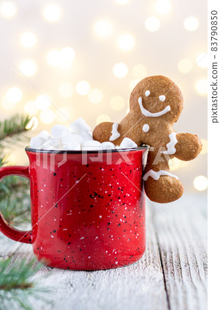 A red cup of winter hot drink with marshmallows and gingerbread man on a white wooden background with bokeh. 83790850