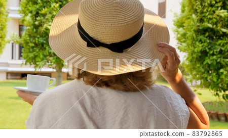 Senora female, age 50-55 in a straw hat holds a cup of hot drink and drinks coffee, sitting in the Senora female, age 50-55 in a straw hat holds a cup of hot drink and drinks coffee, sitting in the 83792865