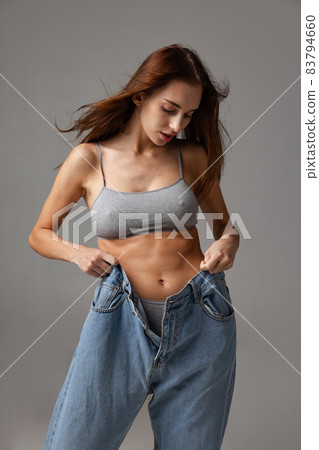 Portrait of young beautiful slim girl in blue jeans jumpersuit posing isolated over gray studio background. Weight Loss concept. Portrait of young beautiful slim girl in blue jeans jumpersuit posing isolated over gray studio background. Weight Loss concept. 83794660