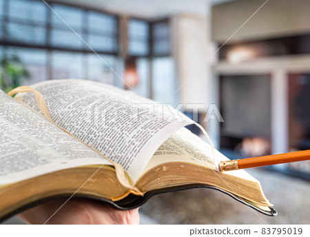 A man holds a Bible with a pencil, against the background of the living room. Reading a book in a cozy atmosphere. 83795019